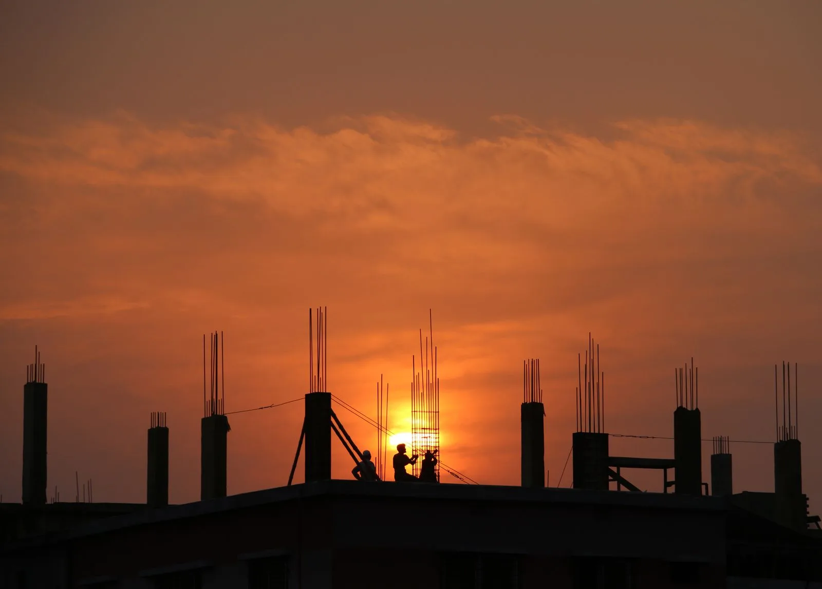 夕暮れの空を背に鉄筋が立ち上がる建設中の建物と作業員のシルエット