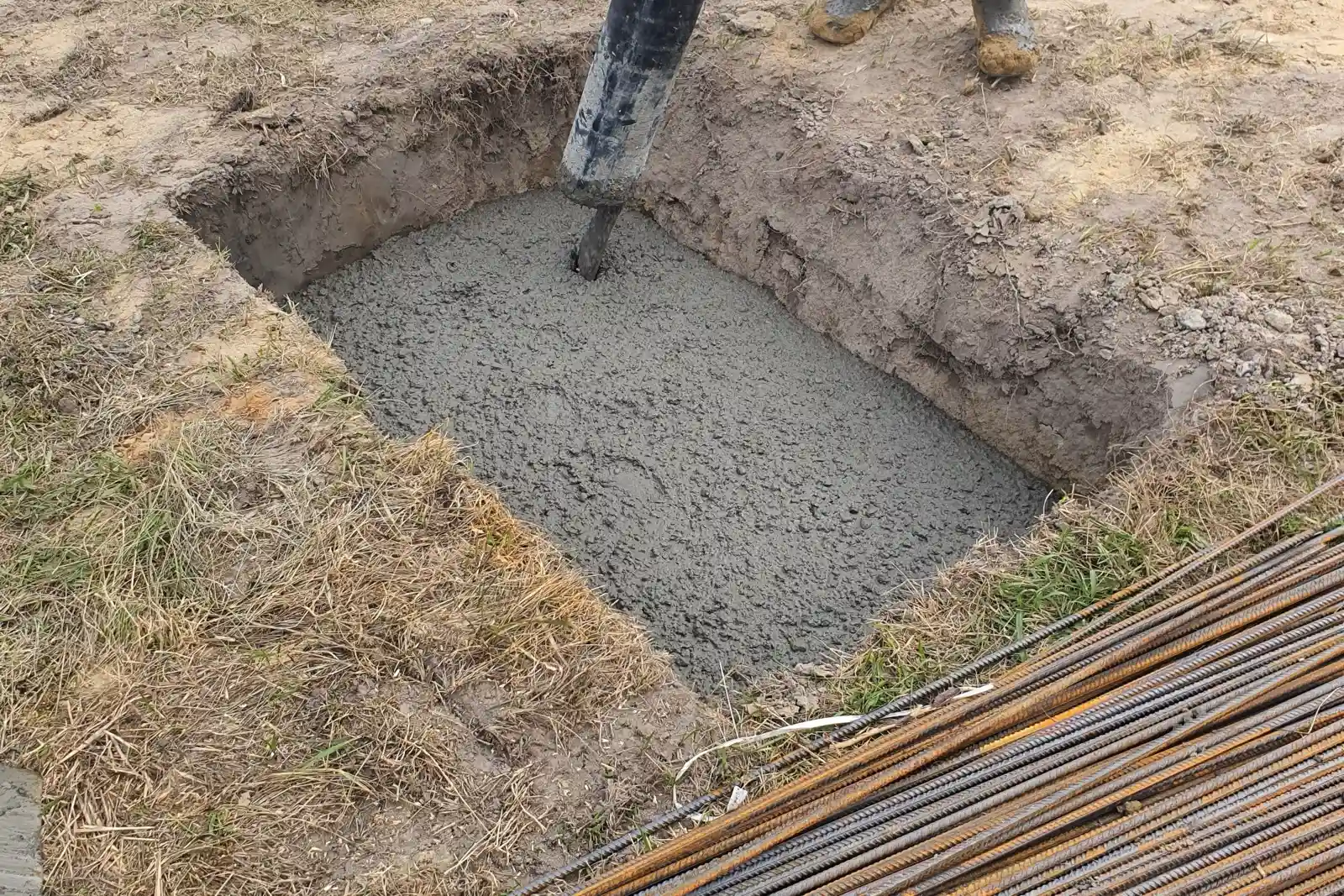 掘削した基礎の穴にホースでコンクリートを流し込む場面と、手前に積まれた鉄筋の束