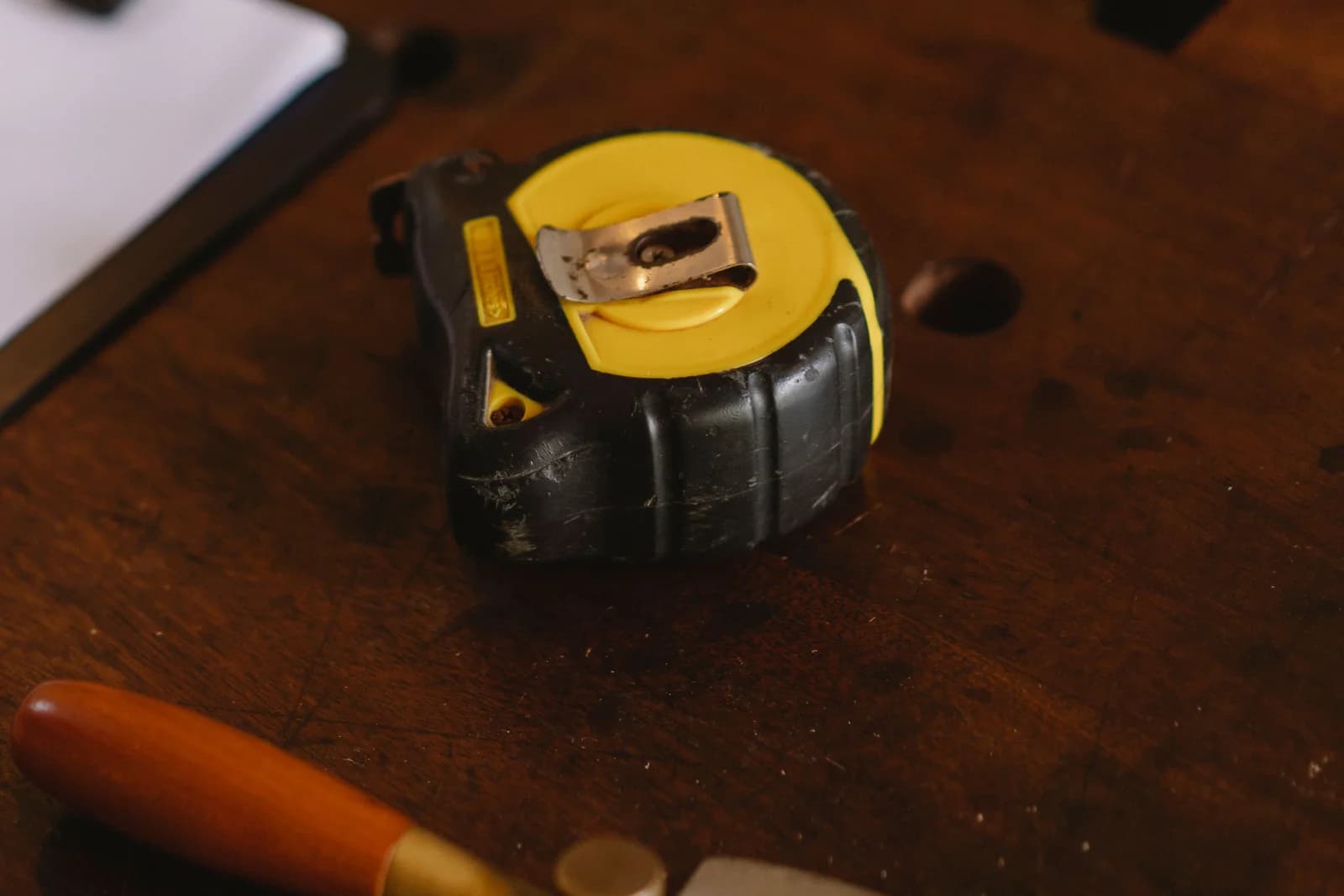 使い込まれた木の作業台に置かれた黒と黄色の巻尺と、木の柄のノミ